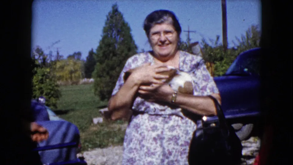 The woman cradles the small dog against her floral dress in a sunny 1960 Wauconda yard with a car and trees behind her.