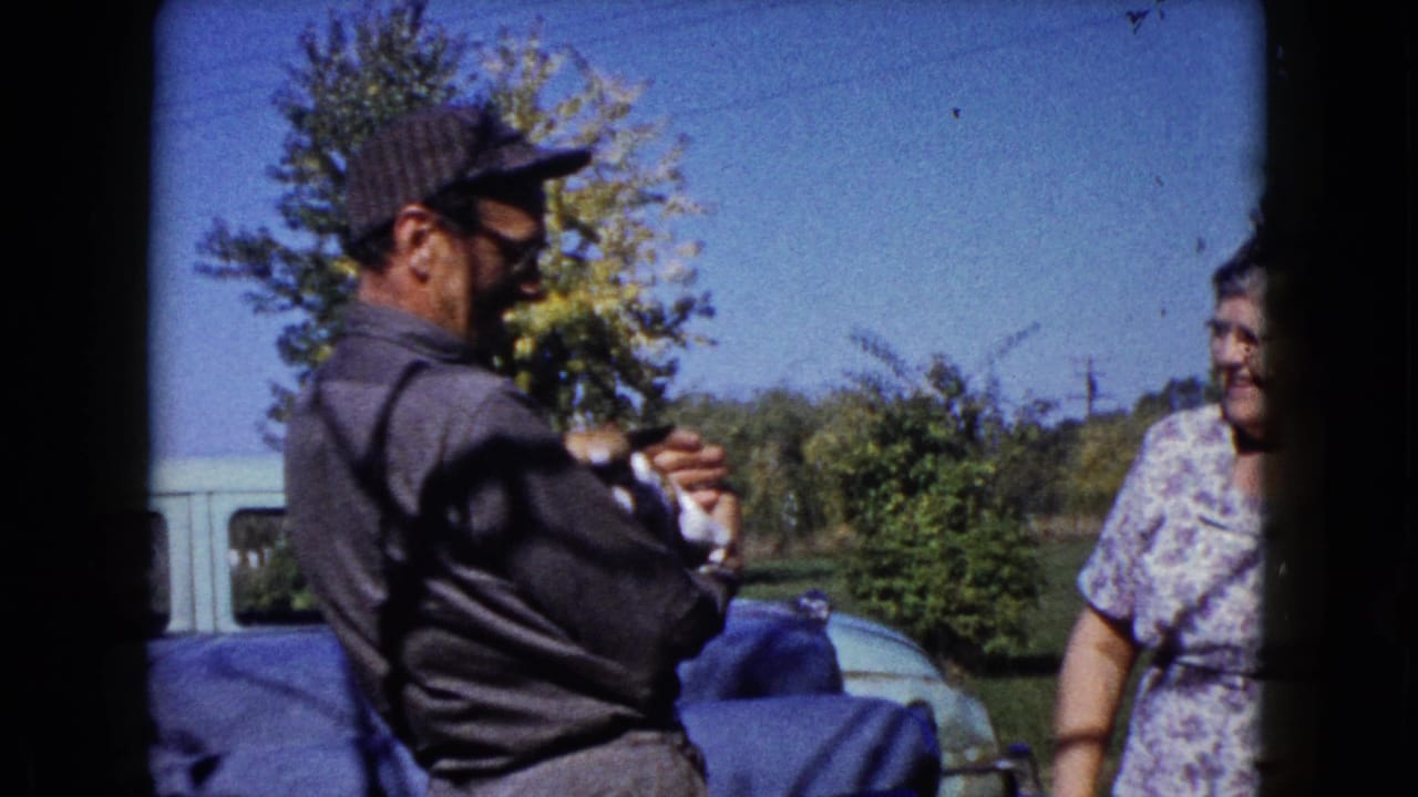 In Wauconda, Illinois circa 1960, a man in glasses and a cap holds a small puppy beside a woman near vehicles and trees.