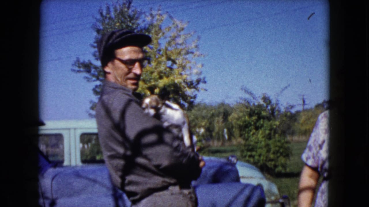 The man looks toward the camera while holding the brown and white puppy, with a pale car, trees, and lawn behind him.