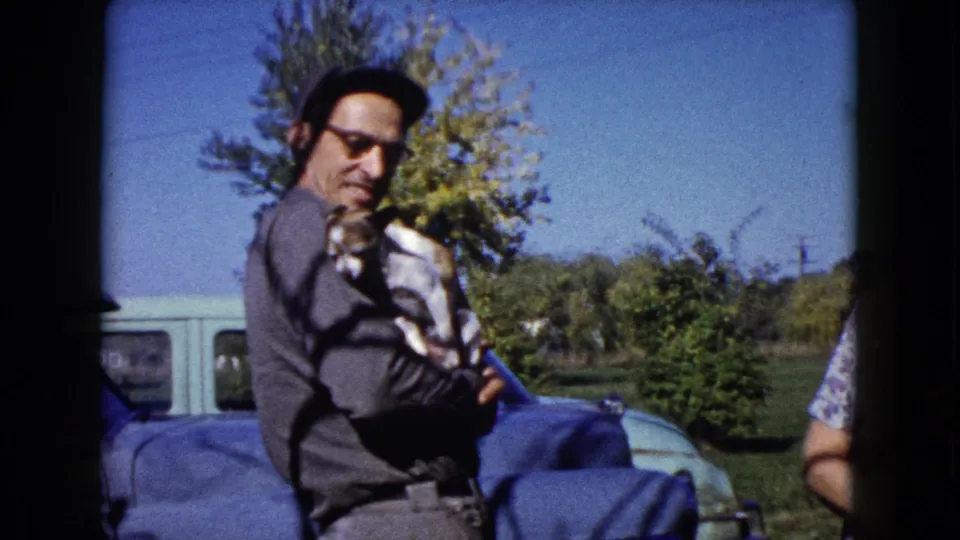 A man in a cap and glasses cradles the small puppy outdoors in the 1960 Wauconda home-movie scene.