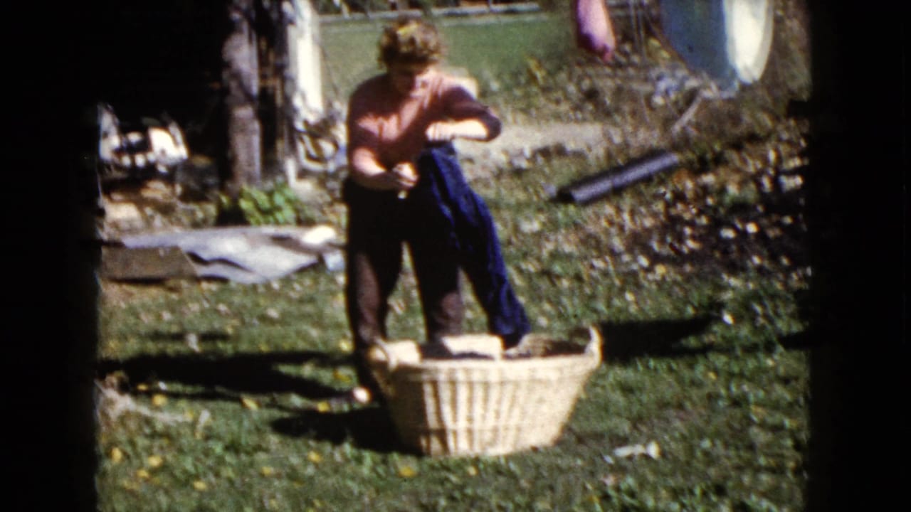 Woman bends over a wicker laundry basket in a sunny backyard near hanging clothes.