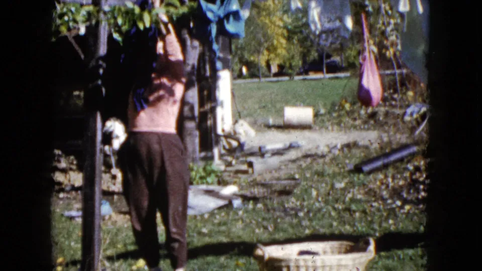 The backyard laundry scene continues as the woman works at the clothesline, with colorful garments and trees visible around her.