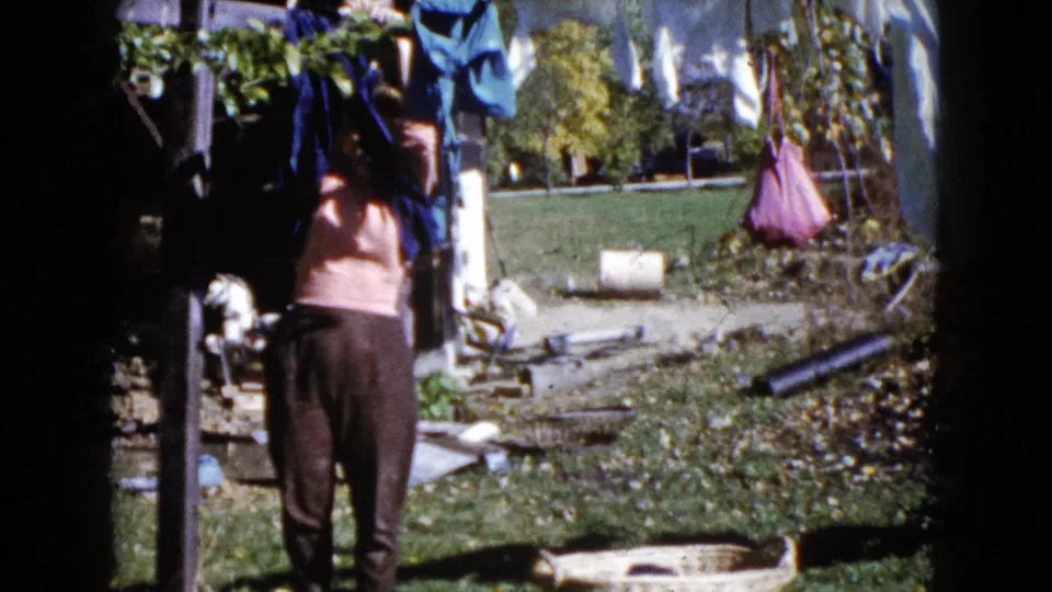 The woman continues hanging or adjusting clothes on a line in a sunlit Wauconda backyard, with grass and scattered yard objects behind her.