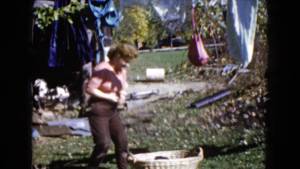 A woman in a pink shirt bends near a wicker basket beneath the clothesline in the 1960 Wauconda backyard scene.