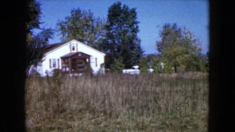 1960 rural Illinois frame showing a white cottage with a covered porch, trees, tall grass, and a light-colored vehicle nearby.