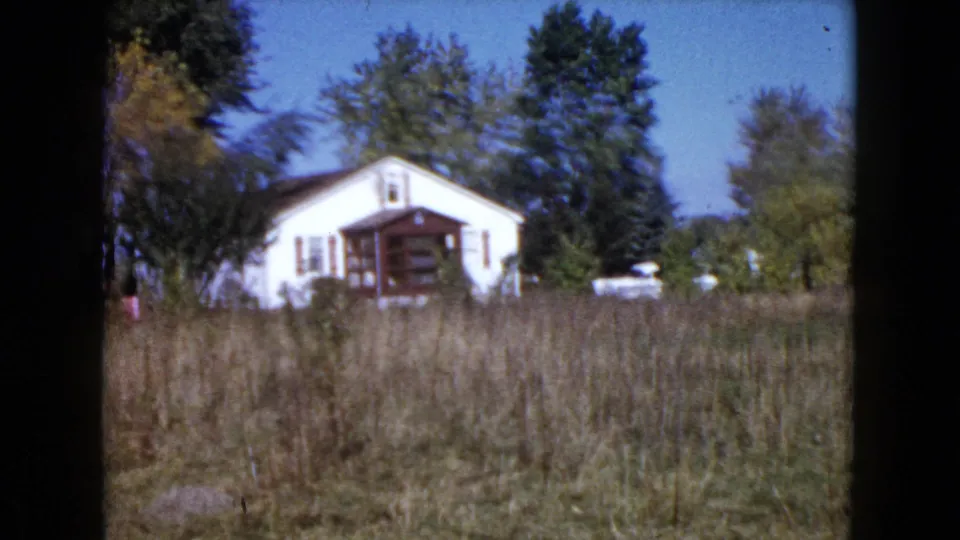Wauconda, Illinois home-movie frame of a white rural house with porch, trees, overgrown field, and a nearby light-colored vehicle.
