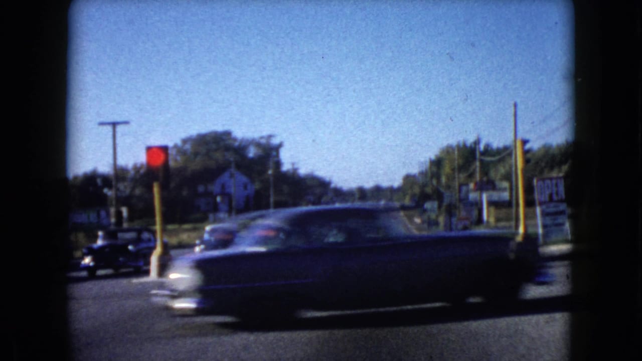 Vintage cars pass through a sunny intersection with a red traffic light and roadside signs.