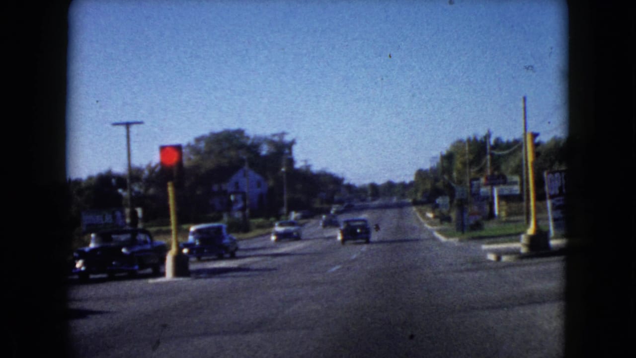 Mid-century cars travel along a Wauconda road in 1960, with a red light, roadside business signs, and trees visible.