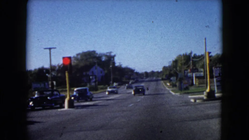 Mid-century cars travel along a Wauconda road in 1960, with a red light, roadside business signs, and trees visible.