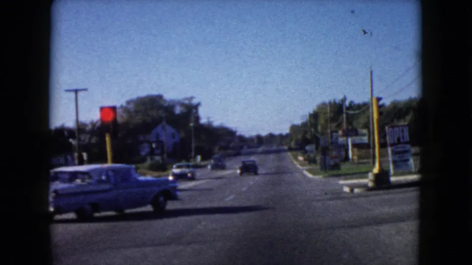 A light-colored 1960-era car crosses a Wauconda intersection, with roadside signs and a quiet road extending behind it.