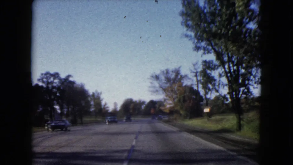 The clip ends on a tree-lined Wauconda road in 1960, with distant cars, lawns, and late-day shadows across the pavement.