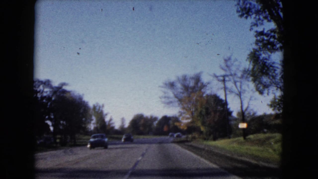 Forward view from a moving car on a tree-lined two-lane road with cars ahead.