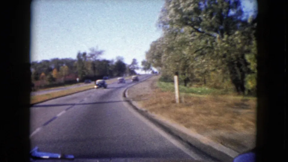 1960 Wauconda, Illinois road-travel frame showing a curving paved road, multiple cars, trees, grassy shoulder, and 8mm film borders.