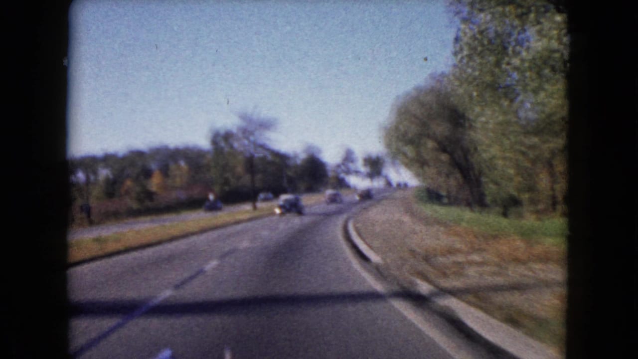 Forward view along a curving two-lane road with trees, grass, and distant cars.
