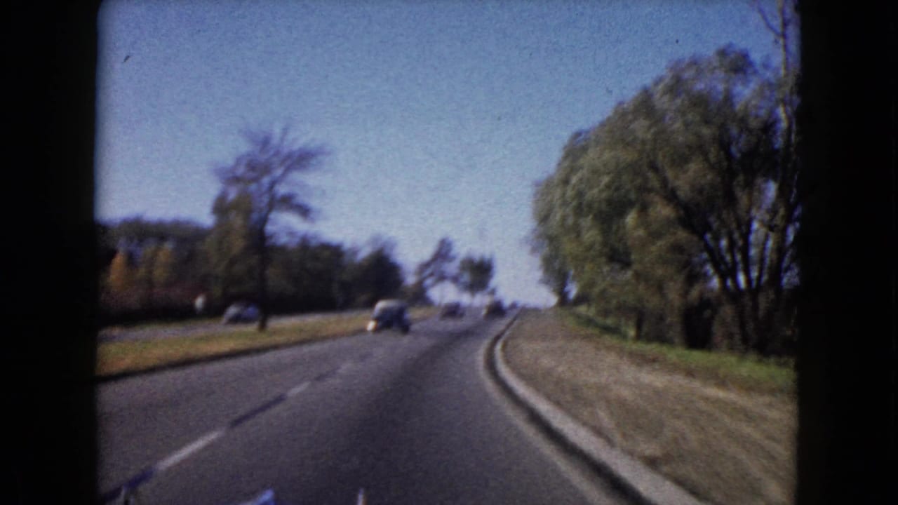 Forward-moving 8mm view of a Wauconda road bending right, with period cars and leafy roadside edges.
