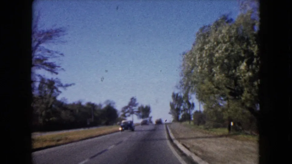 A 1960 home-movie frame shows a slight rise in a Wauconda road, with a distant car and mature roadside trees.