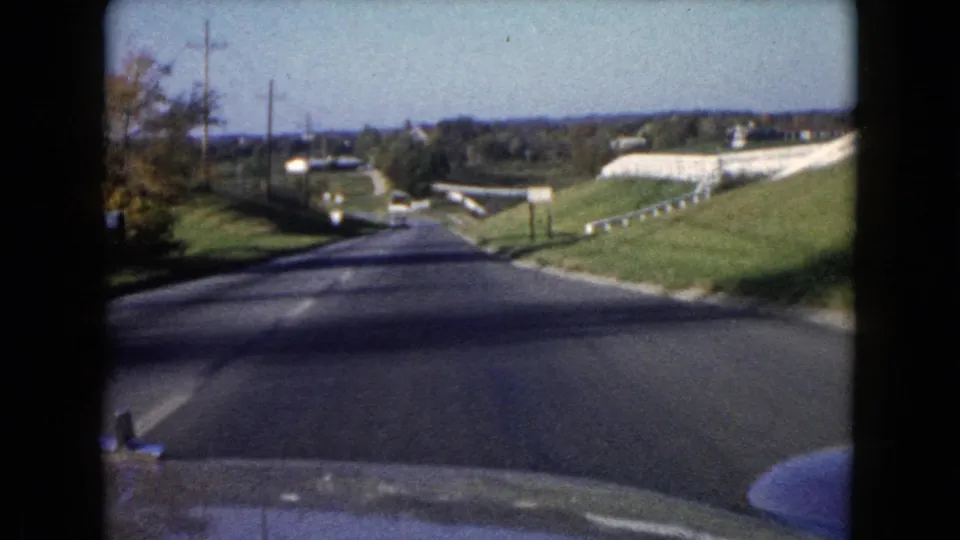 Passenger-view 8mm frame of a Wauconda road descending past cars, utility poles, guardrail, and low roadside buildings.