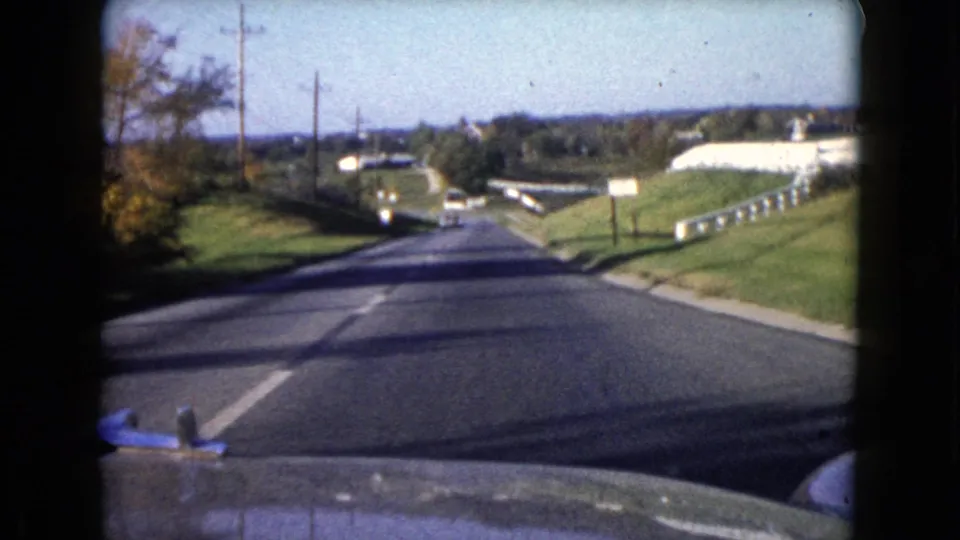 1960 Wauconda, Illinois home-movie view from a moving car on a two-lane road with poles, grass embankments, and distant vehicles.