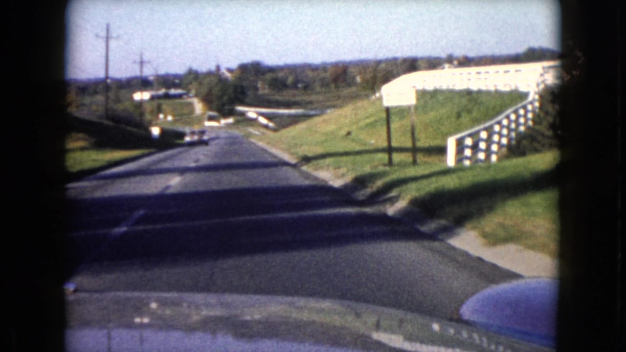 Passenger-view frame from 1960 Wauconda showing a suburban roadway, grassy roadside, utility poles, and a white fence or rail on the right.