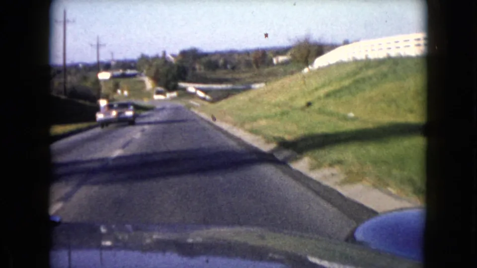 1960 Wauconda road scene from inside a moving vehicle, with an oncoming car, roadside grass, utility poles, and a low building or wall.