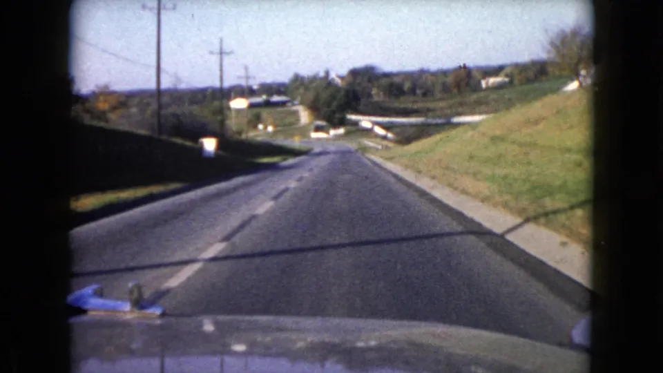End frame of a 1960 Wauconda driving shot, looking over the car hood toward a quiet road with poles, grass, and distant houses or structures.