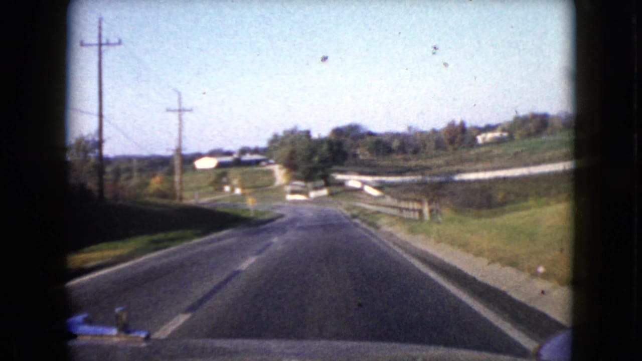 View from a moving car on a two-lane rural road with utility poles, fields, and houses ahead.