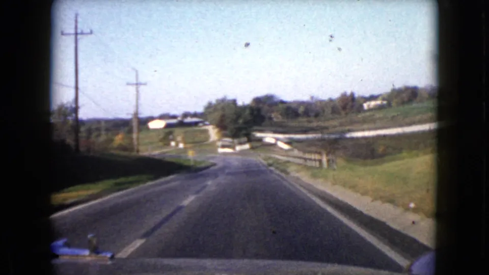 1960 Wauconda, Illinois home-movie view from a car traveling along a rural two-lane road with fields and houses ahead.