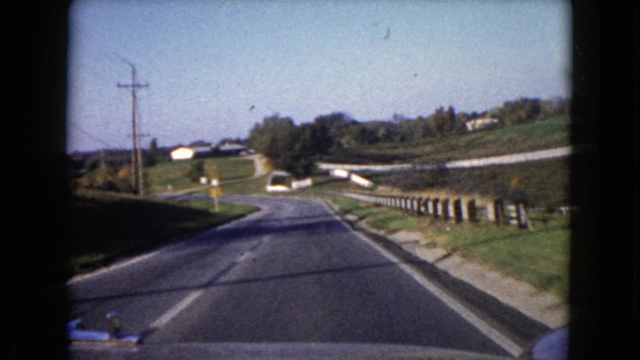 1960 Wauconda, Illinois road scene with a white car ahead on a rural curve, bordered by grass, fencing, and utility poles.