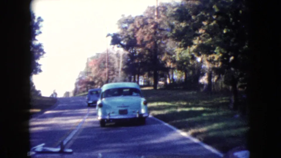 1960 Wauconda, Illinois home-movie frame following a light blue vintage car on a shaded two-lane road.