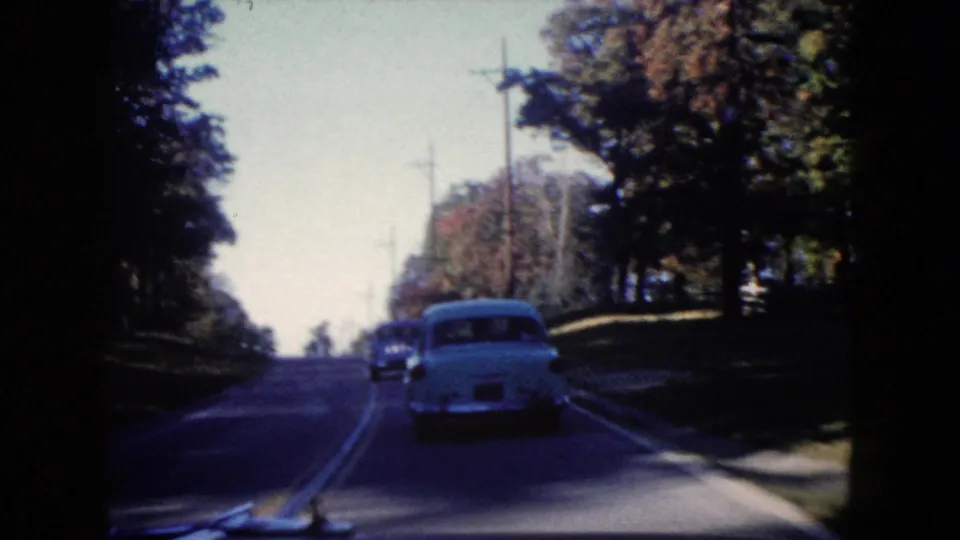 1960 Wauconda, Illinois driving view behind a light blue car on a shaded roadway with trees and utility poles.