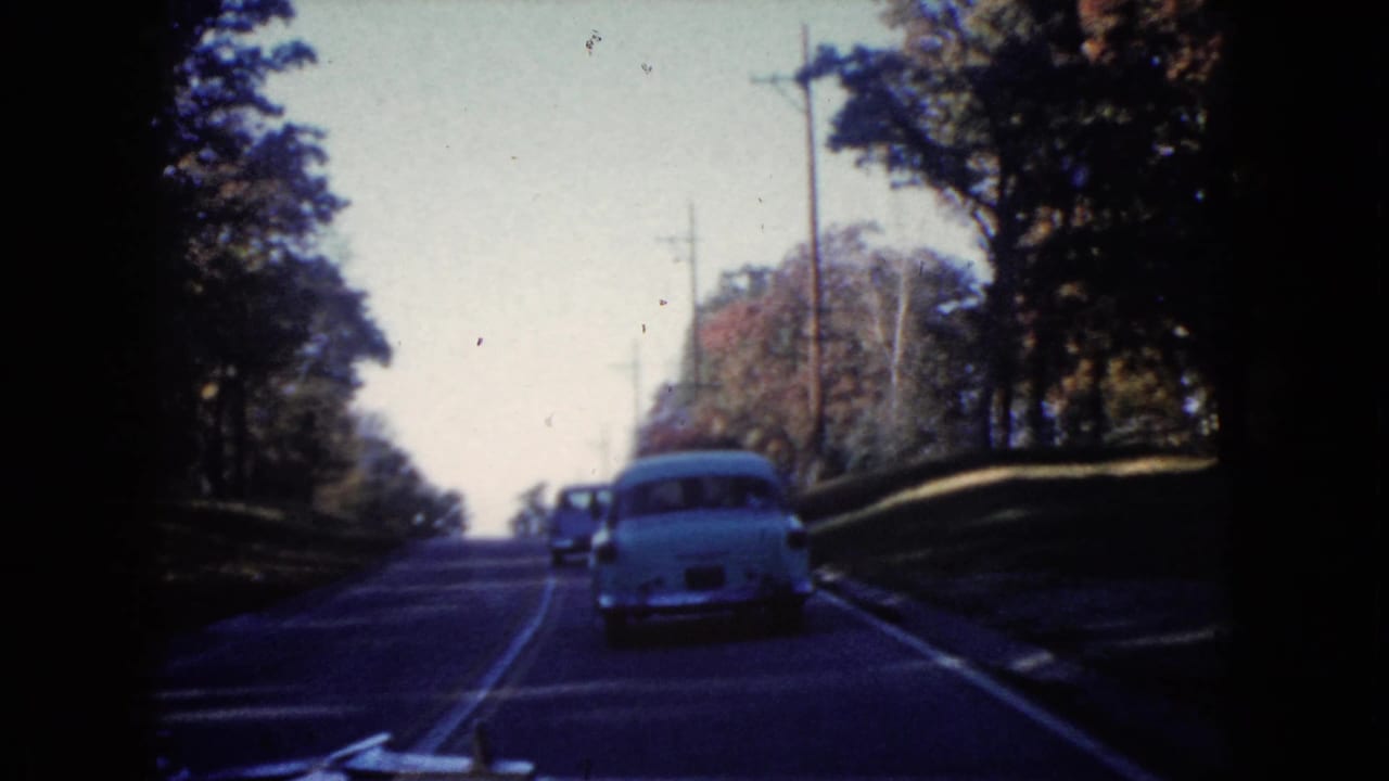 1960 Wauconda home-movie frame showing a white sedan ahead on a gently rising tree-lined road with utility poles.