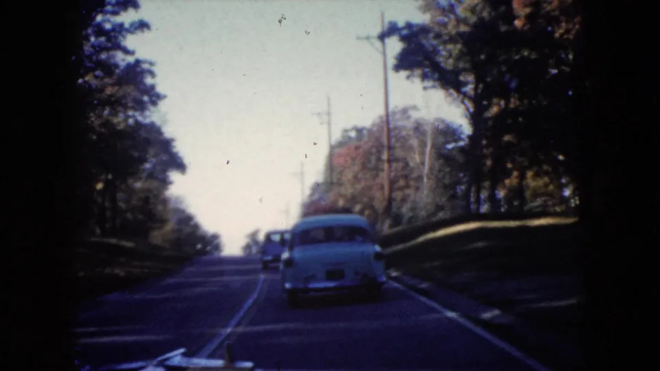 1960 Wauconda home-movie frame showing a white sedan ahead on a gently rising tree-lined road with utility poles.