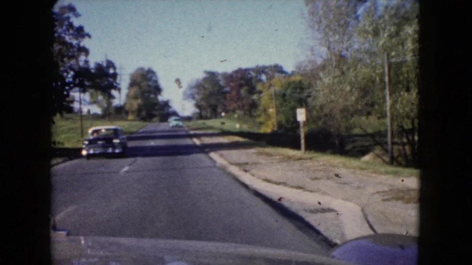 1960 Wauconda road frame showing an oncoming dark sedan, curbside pavement, grassy roadside, trees, and small signs.