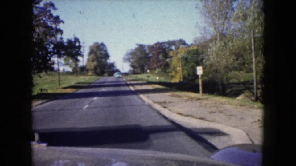 1960 Wauconda home-movie driving frame with a clear road ahead, curb, roadside sign, trees, utility poles, and car hood edge.