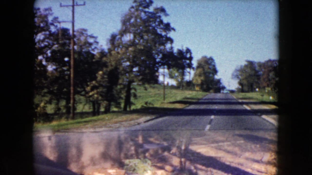 1960 Wauconda, Illinois 8mm frame of a suburban road seen over a car hood, with utility poles, trees, and open grass nearby.