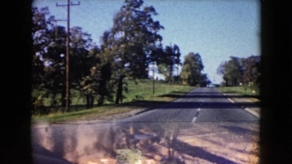 1960 Wauconda, Illinois 8mm frame of a suburban road seen over a car hood, with utility poles, trees, and open grass nearby.