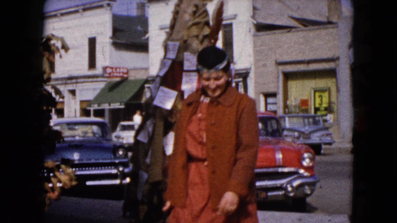 In 1960 Wauconda, Illinois, a woman in a brown coat and black hat walks past decorated street elements and classic parked cars.