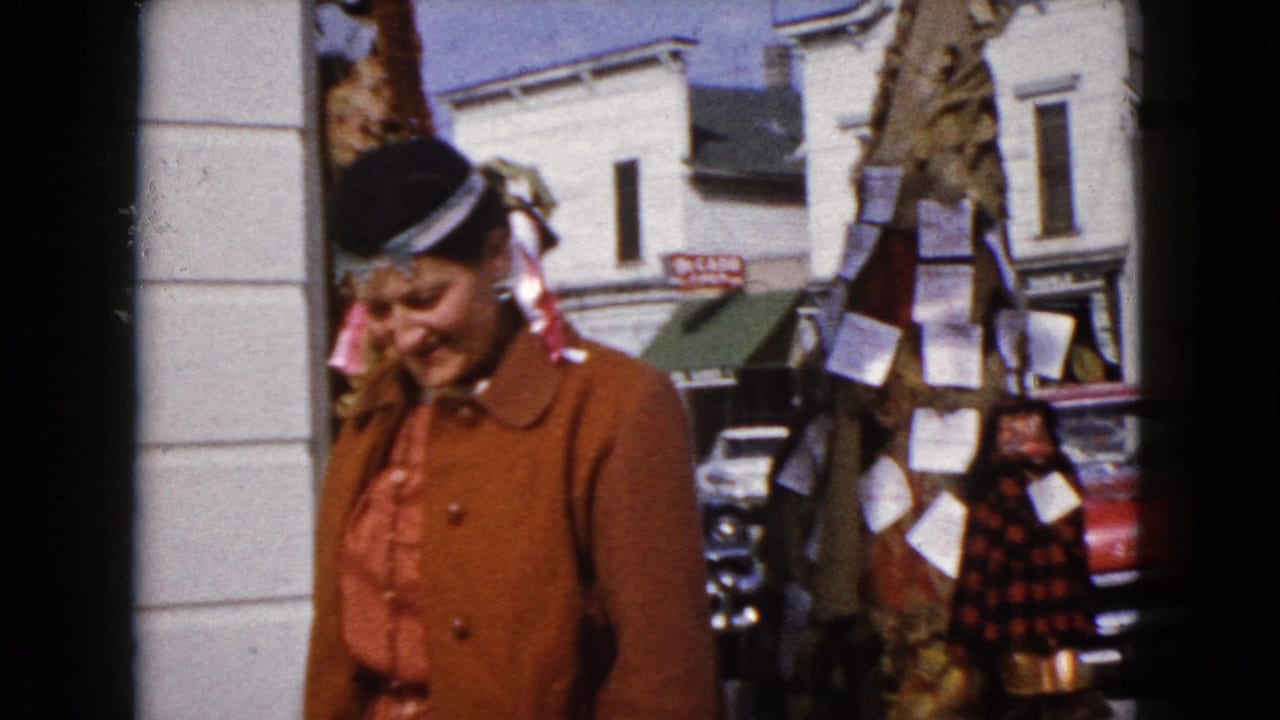 Woman in brown coat passing a storefront column with parked cars behind her.