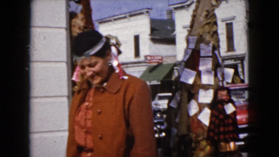 A woman in a brown coat and hat moves past a downtown storefront column in 1960 Wauconda, with classic cars visible behind her.
