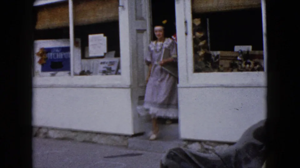 A woman in a gray Native American-style costume stands near a storefront entrance in downtown Wauconda, Illinois, in the 1960 home-movie footage.
