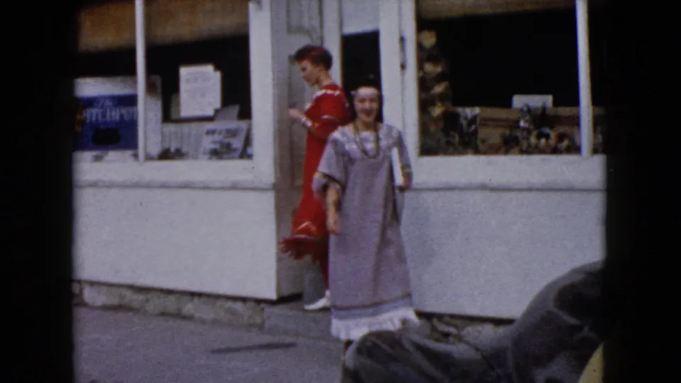 Two women in red and gray Native American-style costumes stand outside a downtown Wauconda storefront, with display windows and signage behind them.