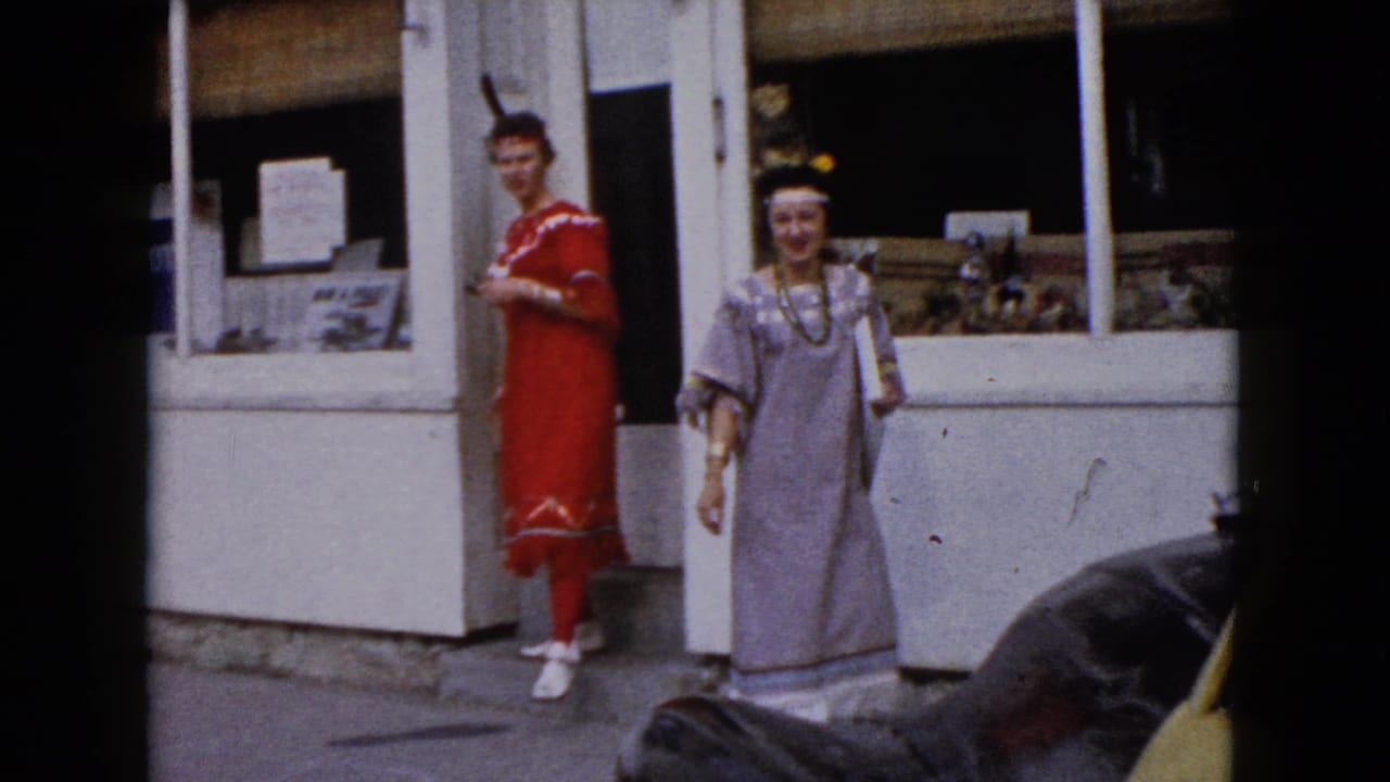 Two women in costume stand outside a white storefront near a parked car.