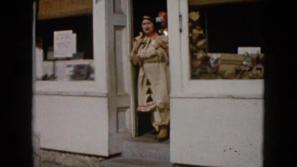 A woman in a patterned Native American-inspired costume emerges from a white storefront doorway in 1960 Wauconda home-movie footage.