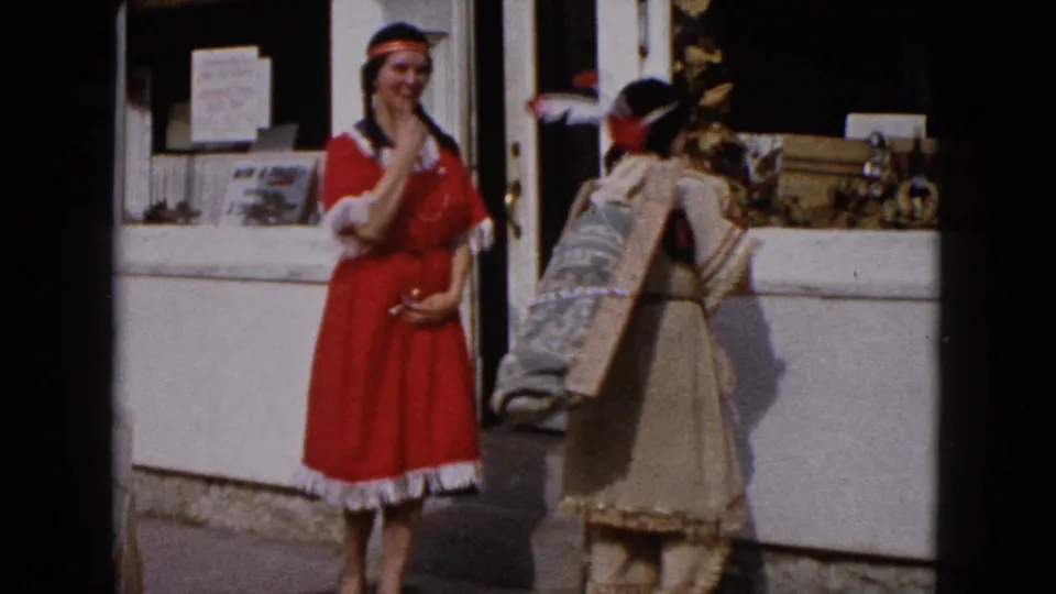 The two costumed women interact outside the Wauconda storefront, with one in a red dress and one carrying a large rectangular prop or sign.