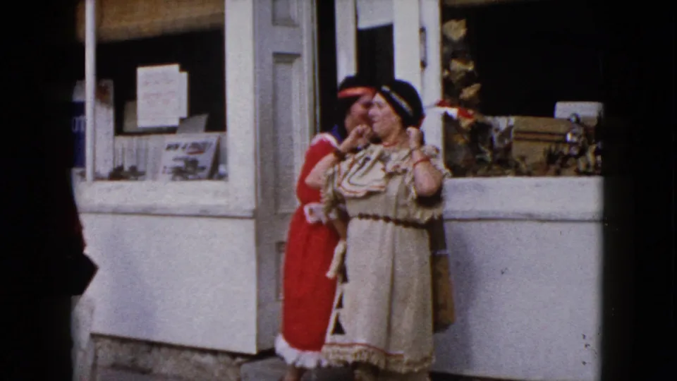 1960 Wauconda frame of a woman in a cream Native American-inspired costume gesturing near a storefront window.