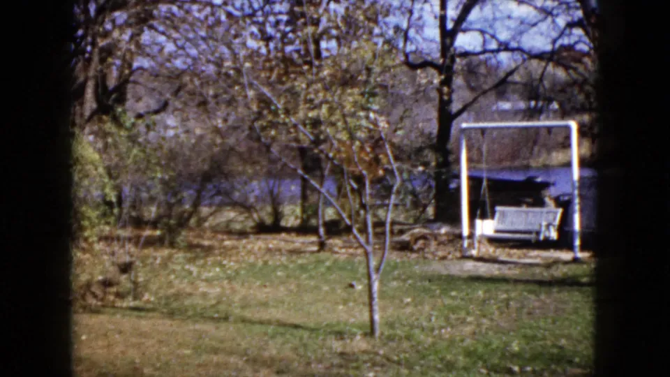 1960 Wauconda home-movie frame showing a spring yard with bare trees, a white swing frame and bench, and water in the background.