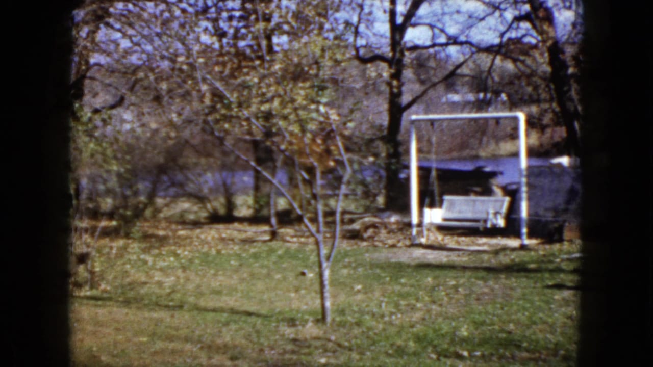 1960 Wauconda yard view holding on the white swing bench, bare trees, grass, fallen leaves, and water behind the property.