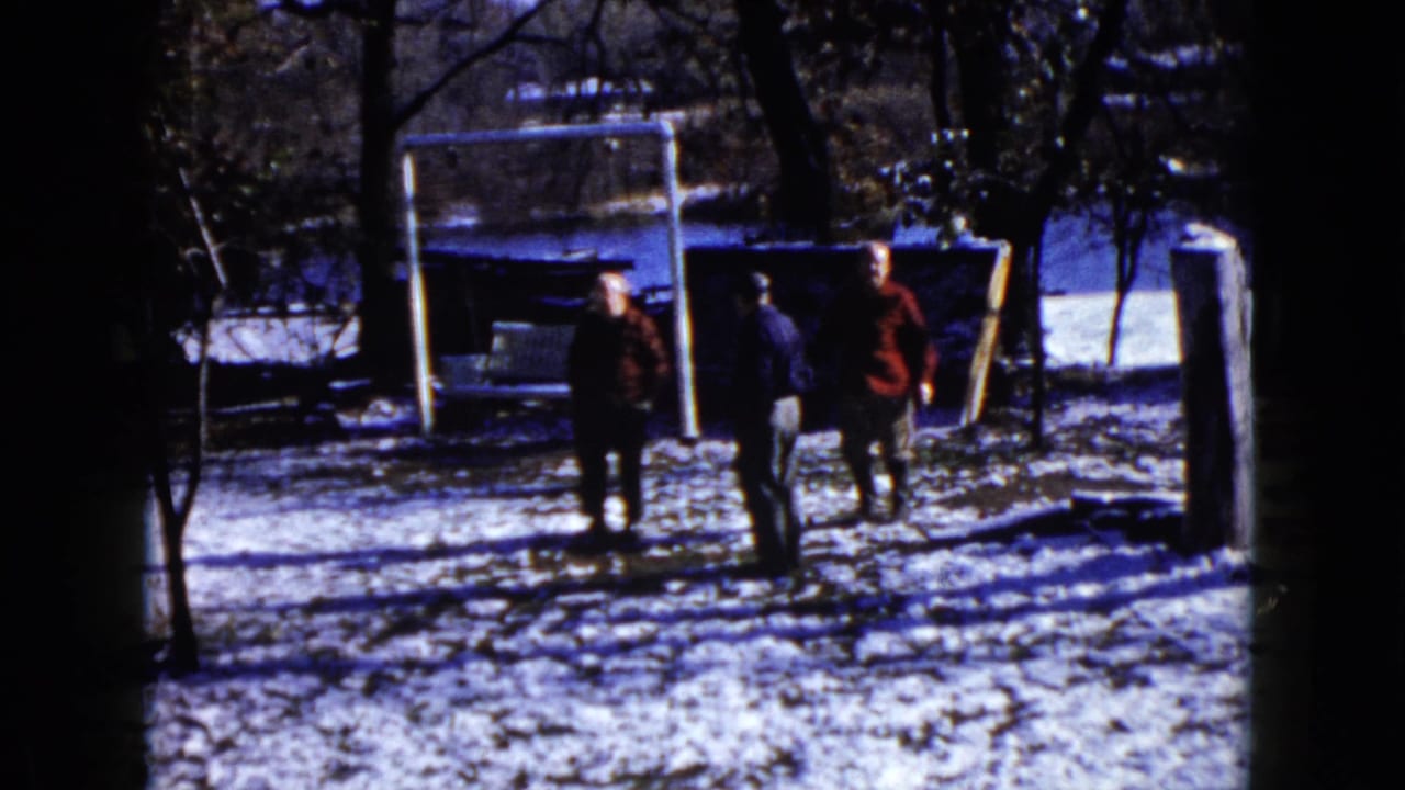 1960 8mm home-movie frame from Wauconda, Illinois, showing three adults in winter coats standing in a snowy yard near a white goal frame.