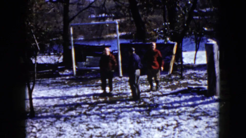 1960 8mm home-movie frame from Wauconda, Illinois, showing three adults in winter coats standing in a snowy yard near a white goal frame.