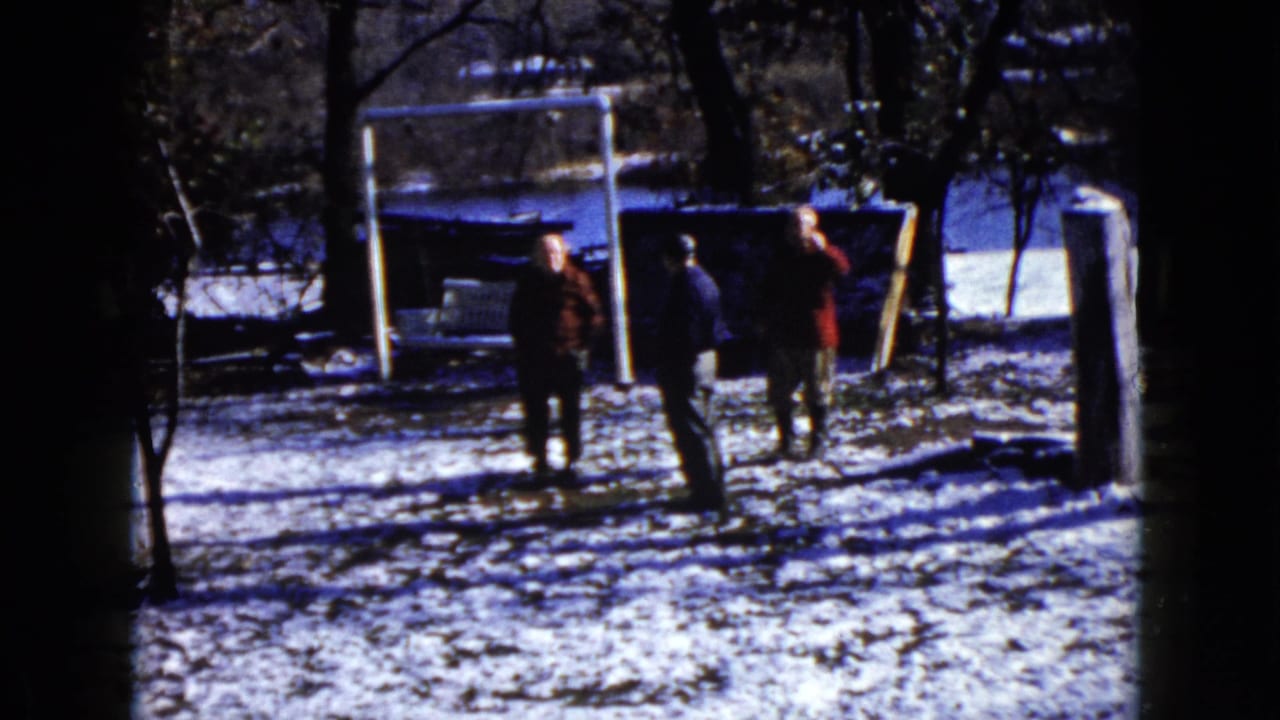 Three adults in coats stand on snow near a goal frame, with one figure beginning to turn.